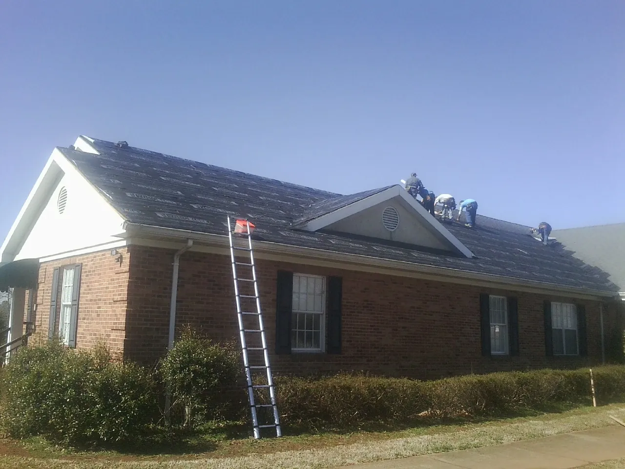 Crew tearing shingles off a brick ranch roof with a ladder set — Emergency Roof Repair work by our crew in Eckert