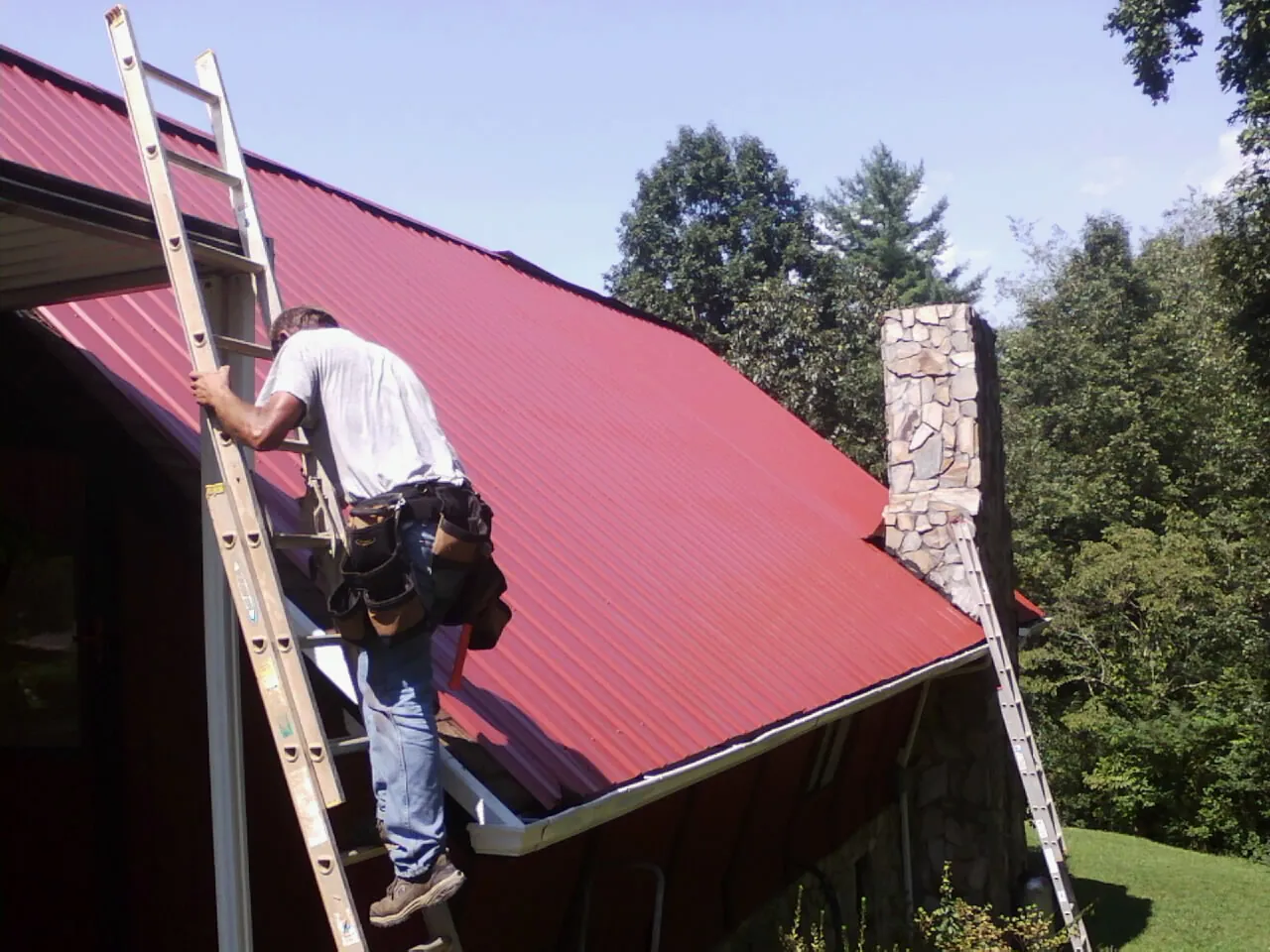 Roofer on a ladder beside a red metal roof and stone chimney — Soffit & Fascia Repair by Roofing Brothers in Eckert